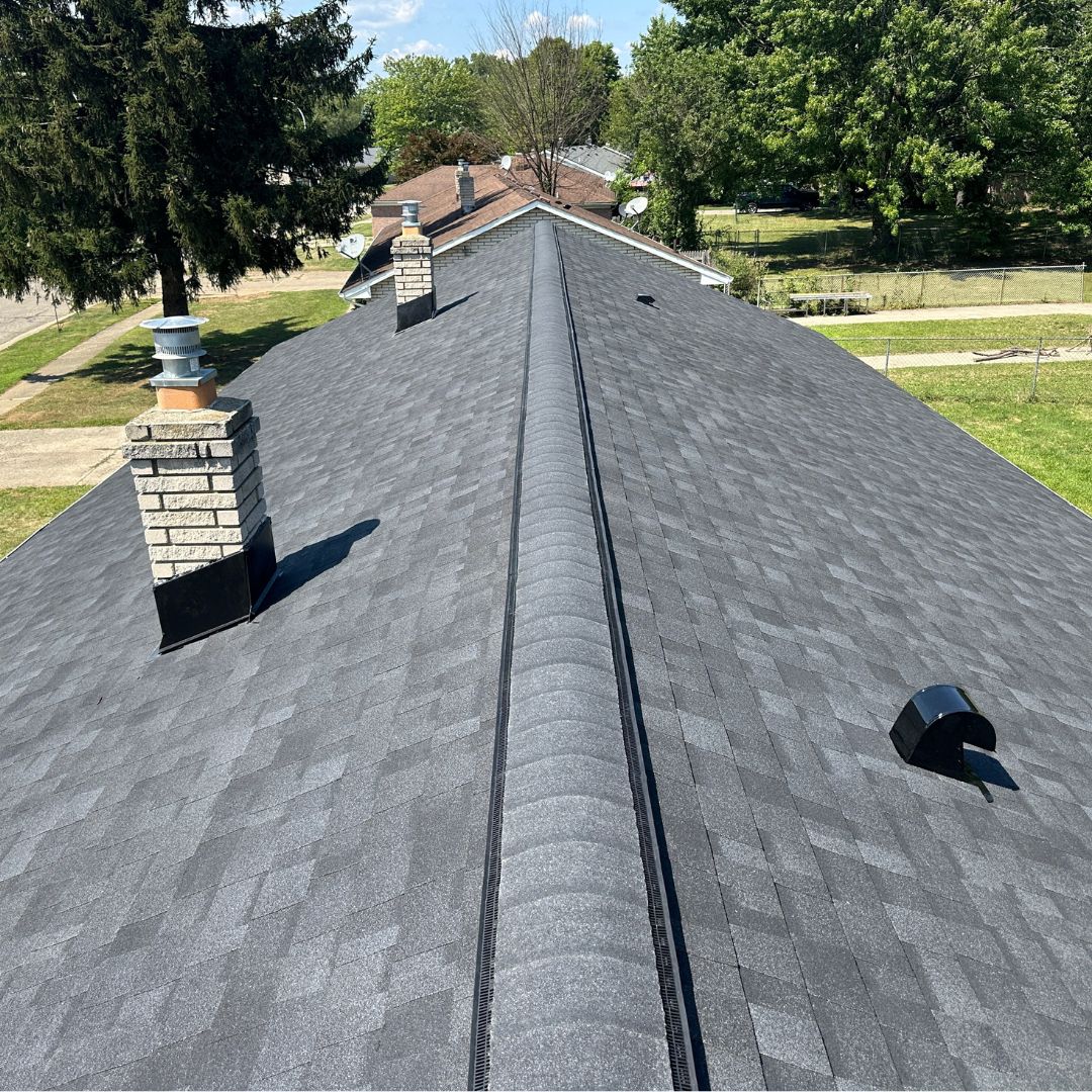 Modern neighborhood roofing with dark gray shingles and stone chimneys under a clear blue sky.